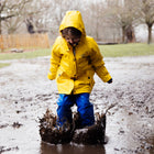 A child in a Puddleflex Waterproof Fleece Lined Jacket Yellow jumps in a muddy puddle, splashing water everywhere.